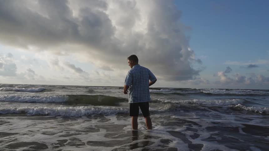 A man wades in ocean waves while fishing on a beach