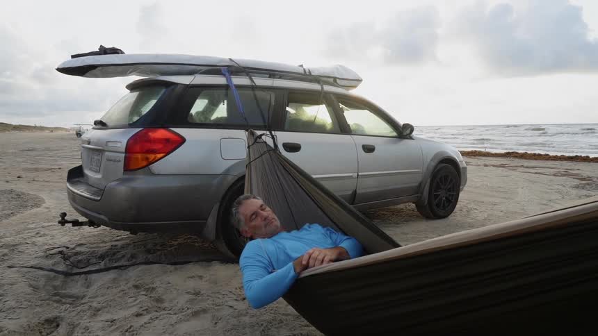 Man sleeps in a hammock that is fastened to his vehicle parked on the beach