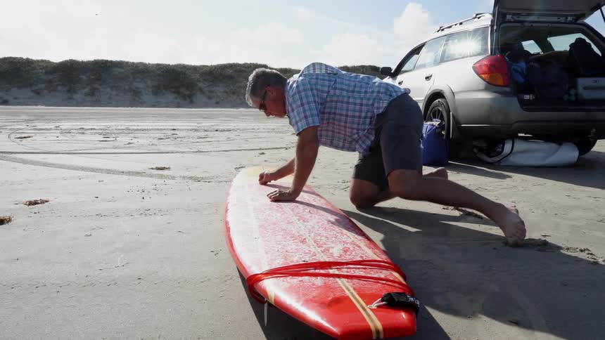 A man waxes his surfboard on the beach near his car