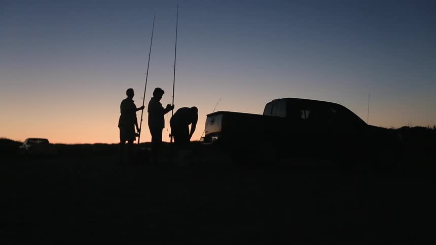 Men stand next to a truck with fishing poles at dusk