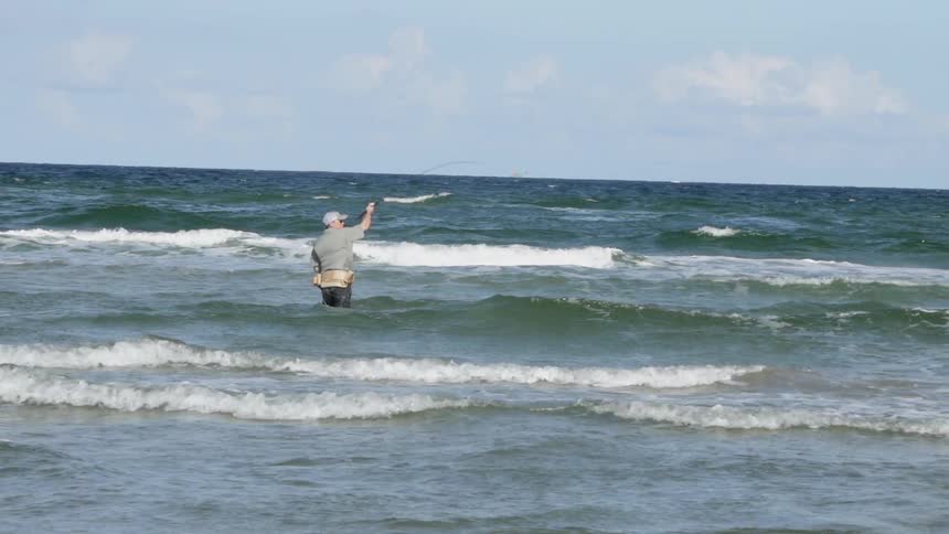 A man wades in ocean waves while fishing on a beach