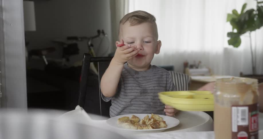 A parent is placing food on the plate of a toddler who is eating inside in daylight