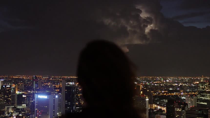 Woman is watching lightning over city at night from balcony