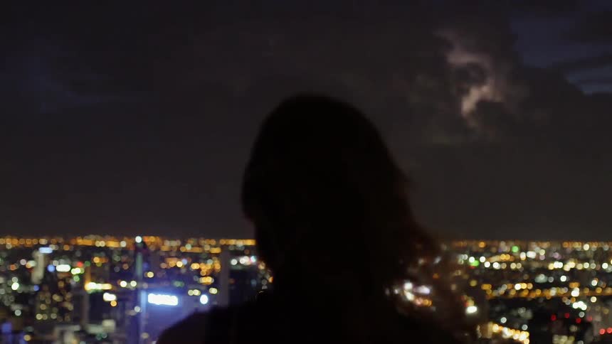 A woman is watching a lightning storm move across a city at night