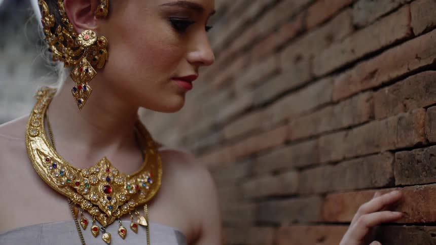 A woman in ornate jewelry touches a brick wall