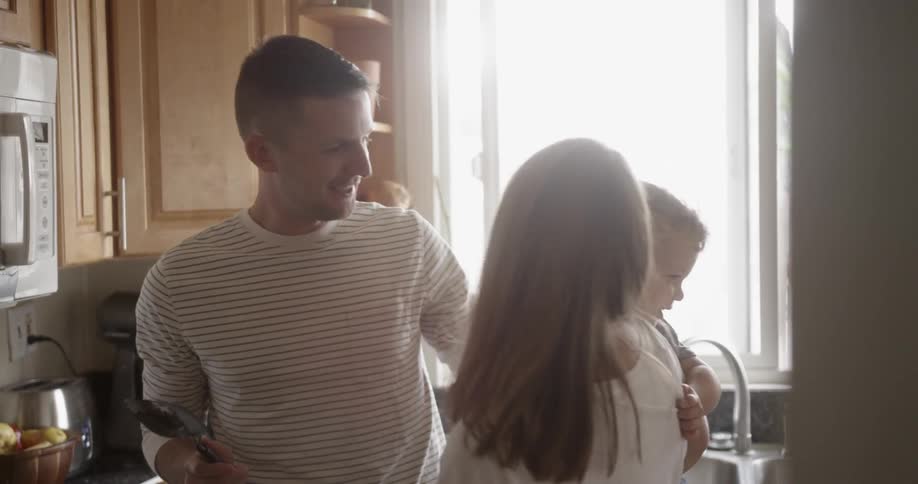 Man kisses a woman holding a baby standing in a kitchen