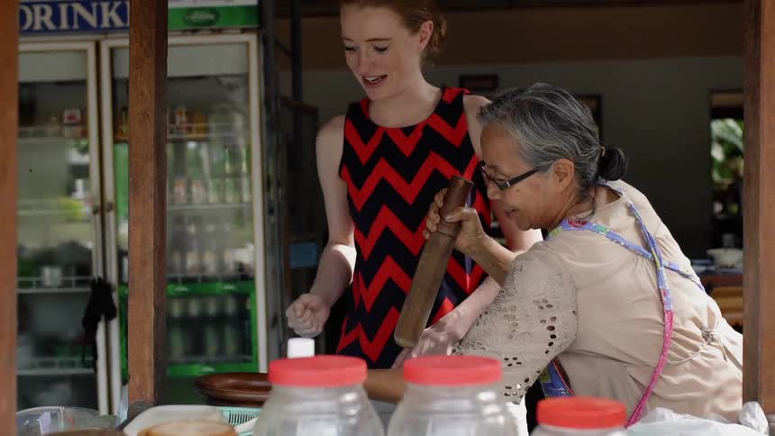 An old asian woman is showing a white woman how to use a pestle outside in daylight