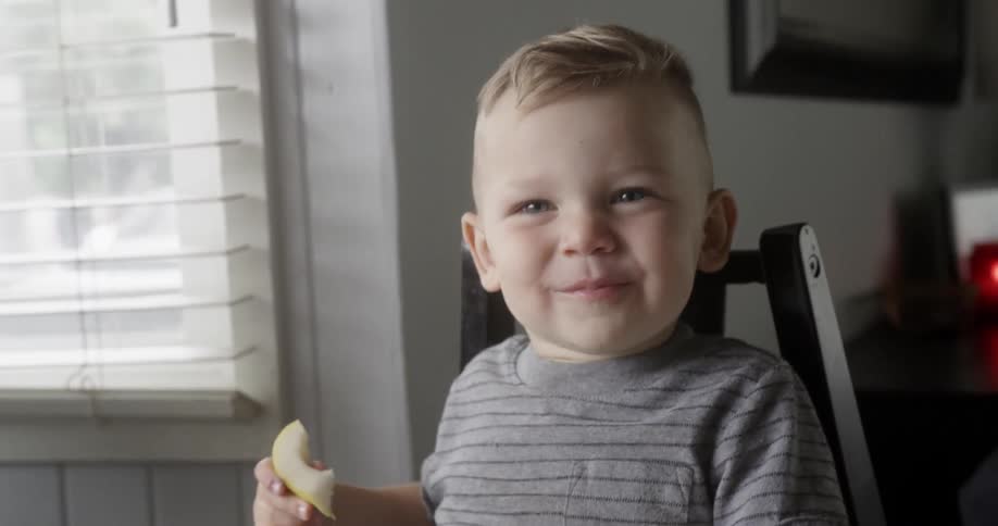 Toddler boy is laughing while eating apple at kitchen table