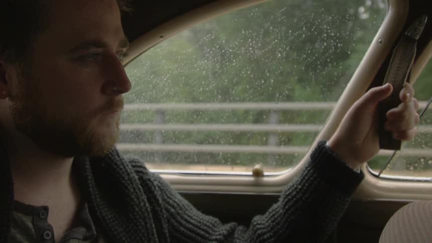 A young, bearded man sitting in a vehicle that has raindrops on the windows and is travelling