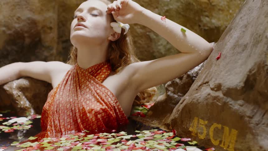 Woman sits in a pool with flower petals in it and a small waterfall splashing behind