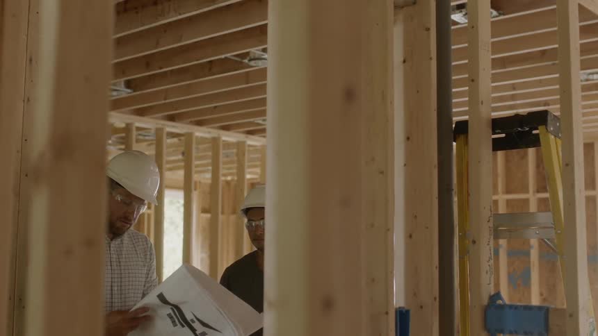 Men holding plans walk through a framed house pointing and talking