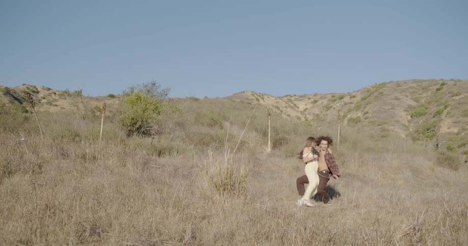 A man hugs a woman running in the grass and they both fall to the ground