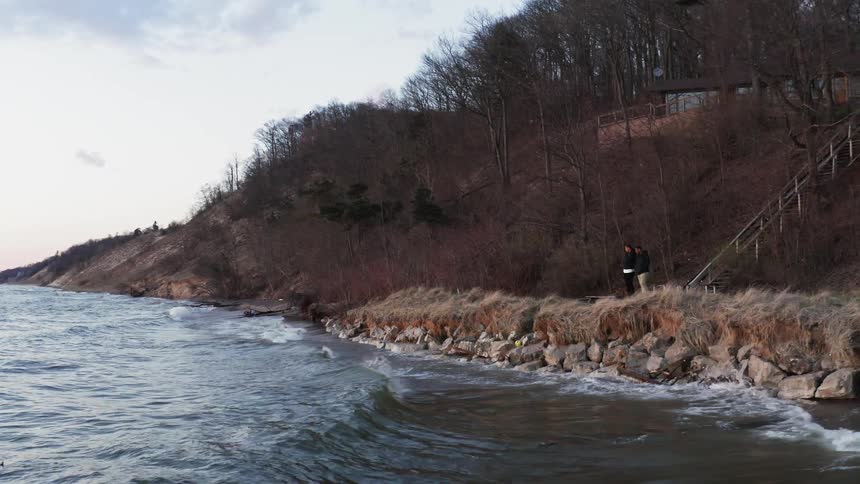 Two people stand on the shore as waves crash