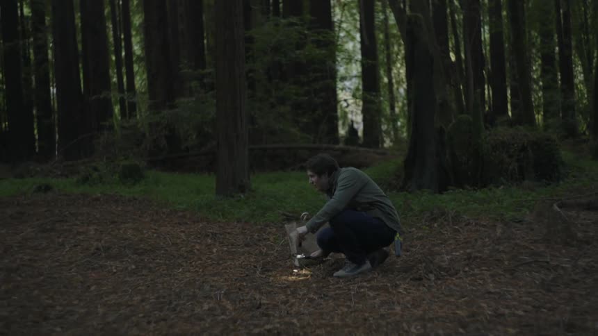 A man using a flashlight and magnifying glass to look at something on the ground in a wooded area