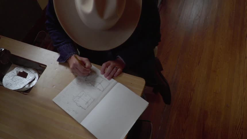 Man wearing cowboy hat is drawing in sketch book at desk