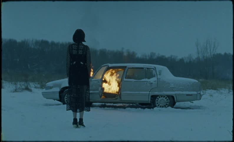 A woman stands in the snow watching a car burn
