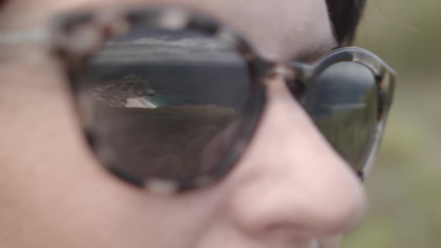 A closeup view of a person's face, with the ocean and shoreline visible in the person's sunglasses