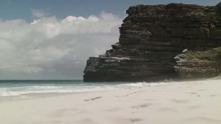 Waves crashing onto a beach next to a cliff on a semi-cloudy day