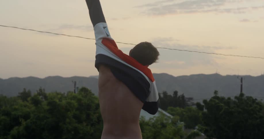 A man pulls on a shirt outside at sunset