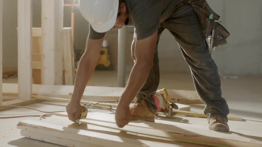 Man wearing a hard hat uses a tape measure to measure the lumber
