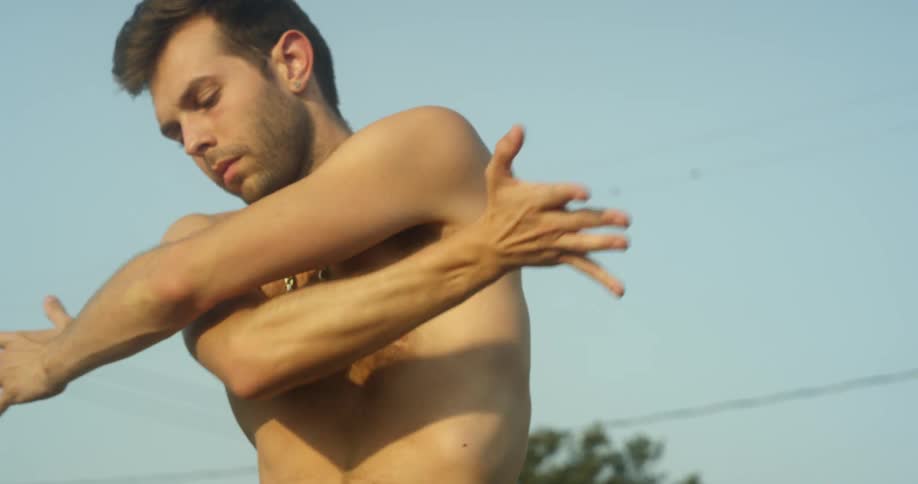 Shirtless man is stretching his arms outside on clear day