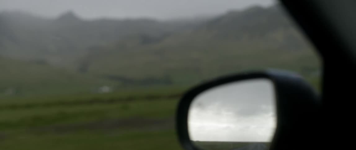 Clouds and road are visible in a car's side mirror as it passes fields