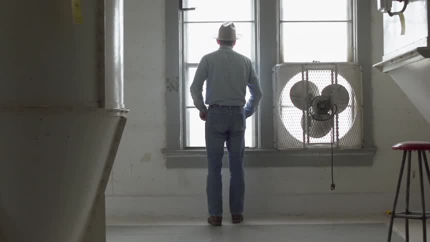 Man wearing a cowboy hat looks out a window standing next to a fan