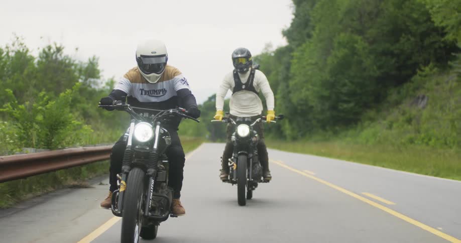 Two men are standing on and riding motorcycles on country road