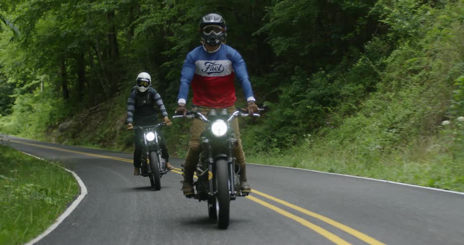 Two people are standing up while riding motorcycles on forest highway