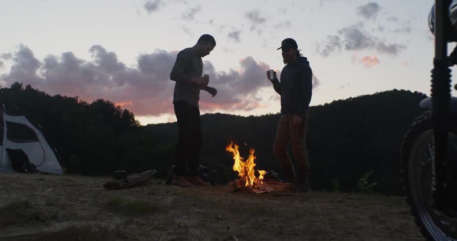 Two men toast their drinks as they stand near a bonfire at a campsite