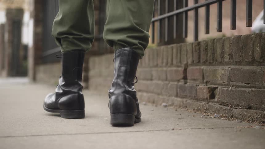 A person wearing boots walks on a sidewalk past a fence