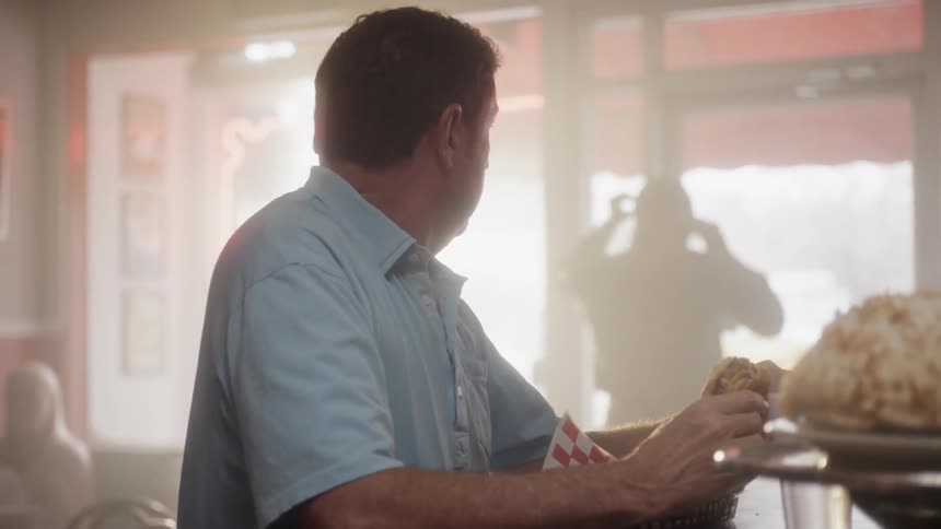 A man sits at a counter eating a burger as a man exits the diner