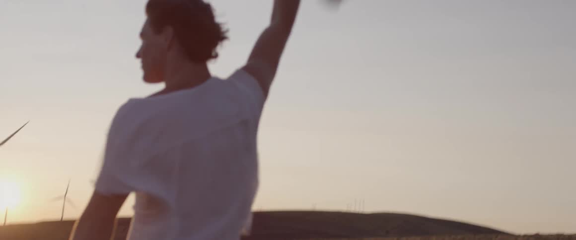 A man is holding his jacket in the air outside at a wind farm at sunset
