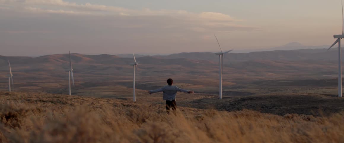 Man stands in a field with outstretched arms near three windmills