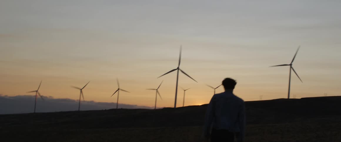 A man walks toward wind turbines at sunset