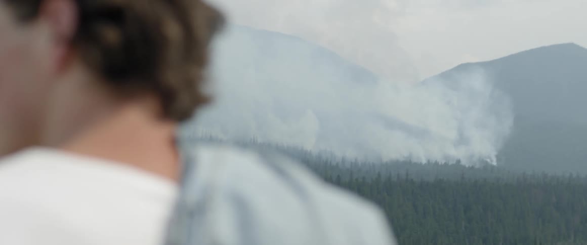 Man stands outside on a hill overlooking trees and smoke