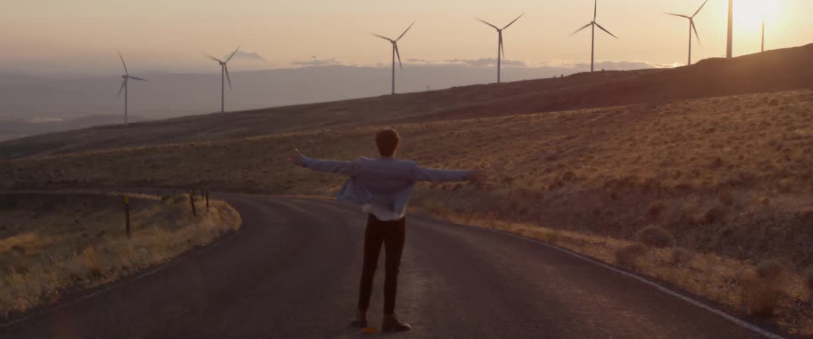 Man holds his arms out wide as he watches the wind turbines