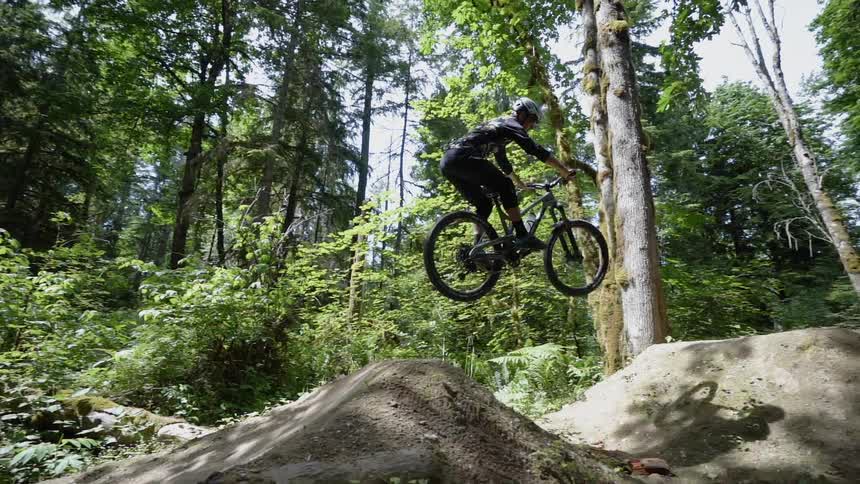 Men ride bicycles over a ramp in the forest
