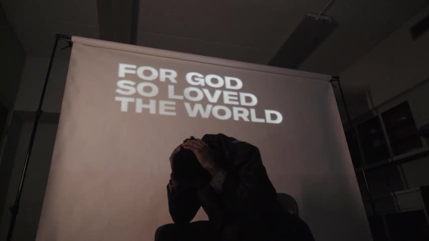 A man sits with bowed head in front of a screen where words are projected