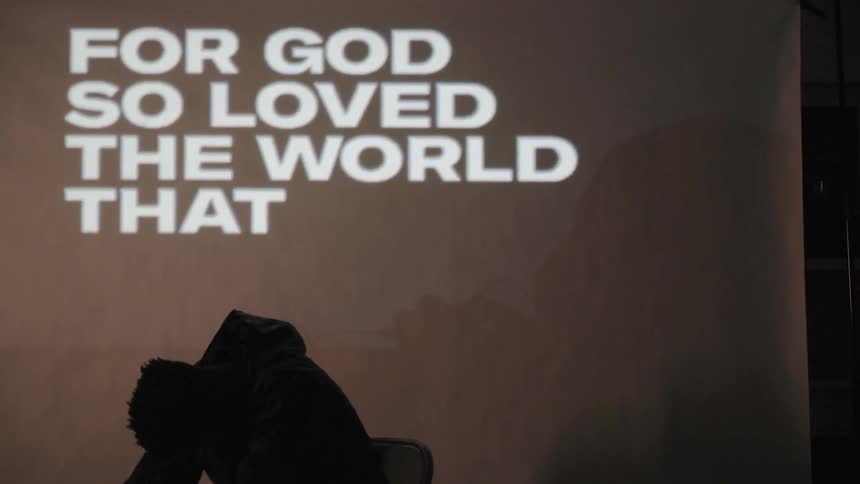 A man sitting in front of a projection screen bows his head as words flash