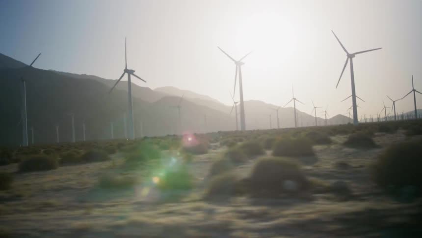 Sun shines over a bushy field and hills with many windmills spinning