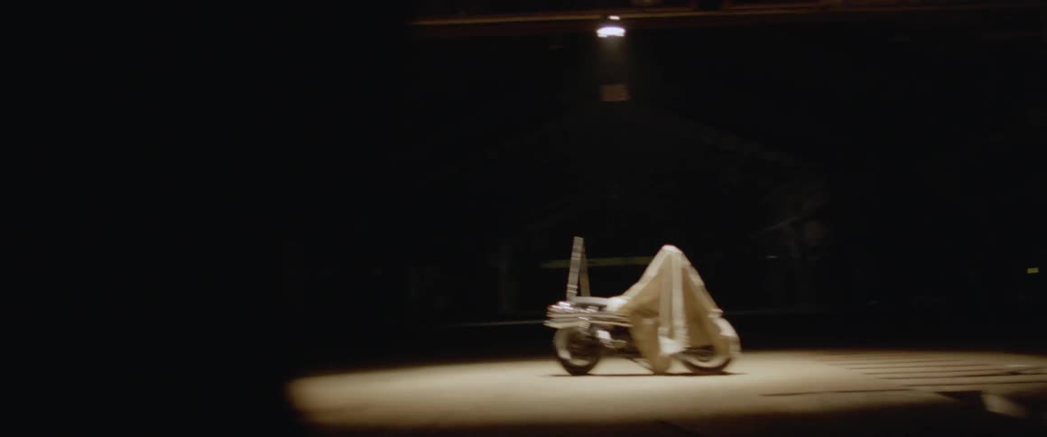 A man walks towards a motorcycle in a dark warehouse