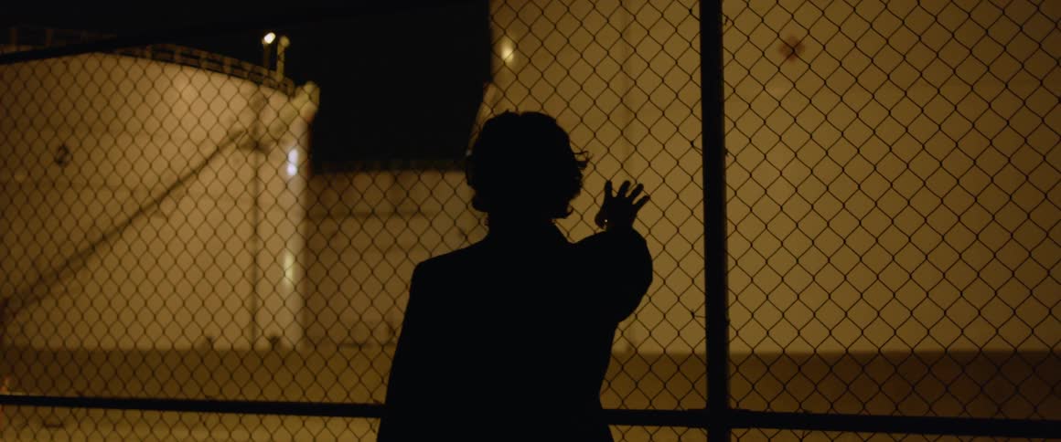 A man is touching a fence outside a refining facility at night