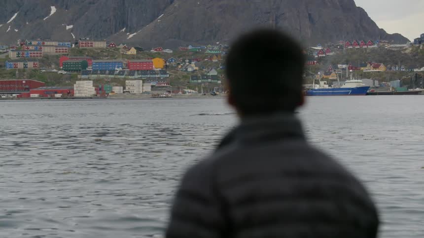 A man in a jacket looks across a bay toward a colorful village with snowy cliffs in the background