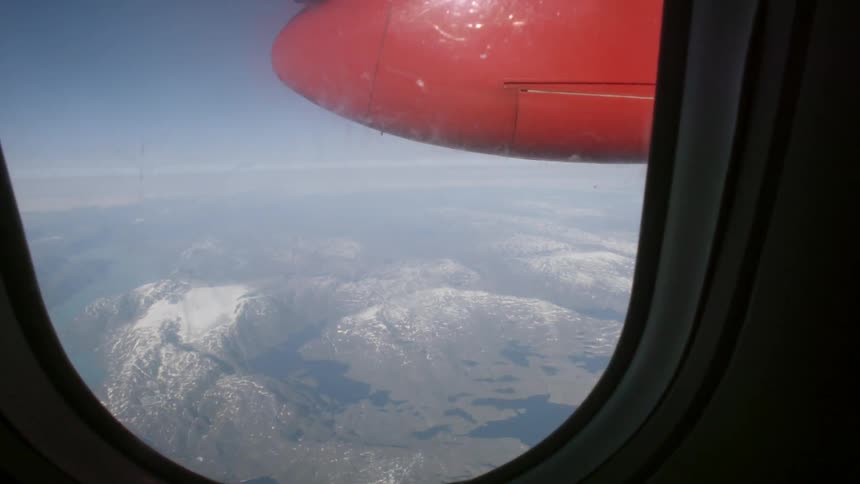 An airplane window overlooks a mountain range
