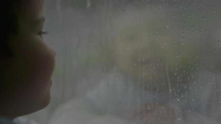 A boy looks out a window as it rains outside