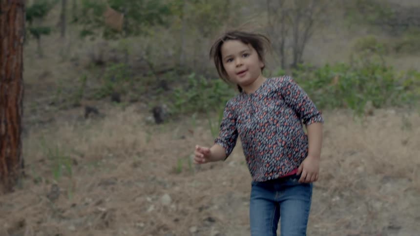 A young, brunette girl bouncing on something next to a hillside that is dotted with trees and brush