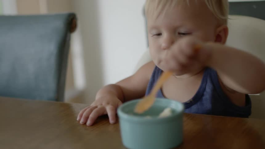 A baby girl sits in a high-chair at a dinner table eating from a small bowl using a spoon
