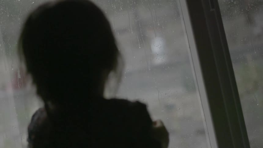 A young girl watches the rain through a window from inside