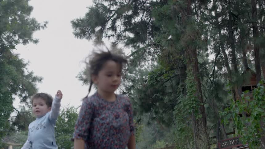 Two small, brunette children bouncing on something in a wooded area